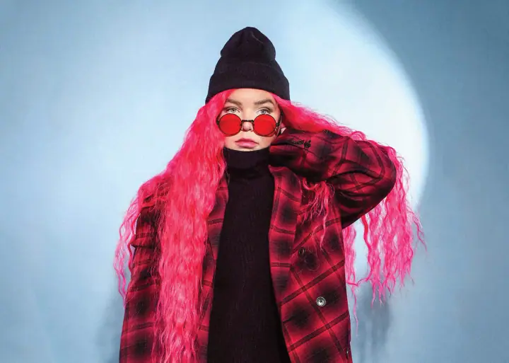 Person with long pink wavy hair, red round sunglasses, and a black beanie in a red plaid coat against a blue backdrop.
