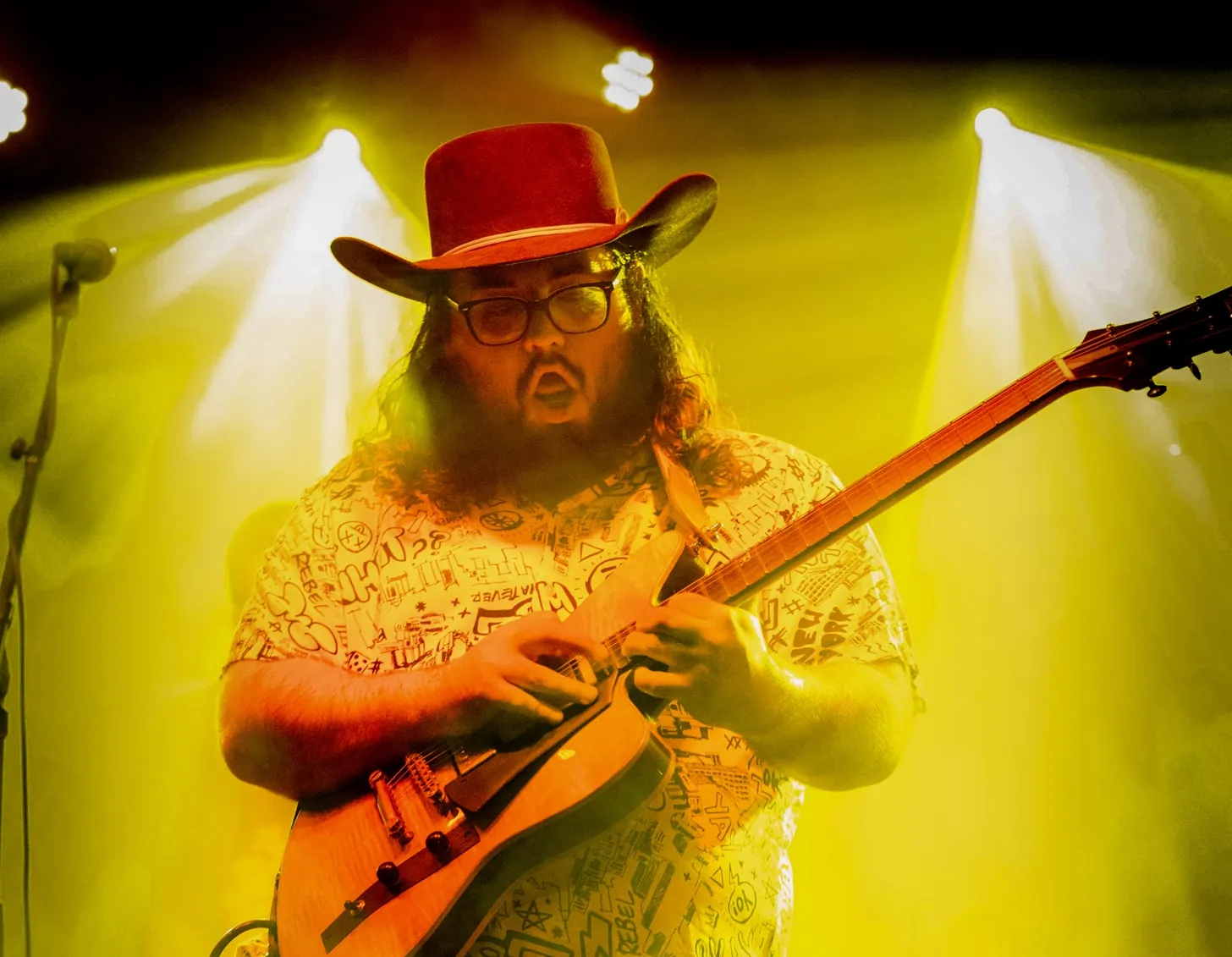 A bearded man in a red cowboy hat and glasses plays an electric guitar under yellow stage lights.