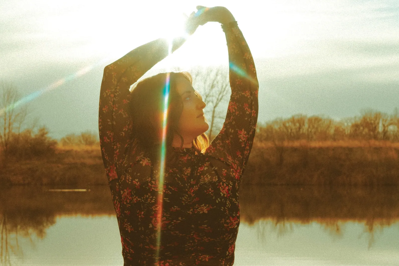 A woman in a floral dress raises her arms overhead in golden sunlight beside a calm lake with bare trees.
