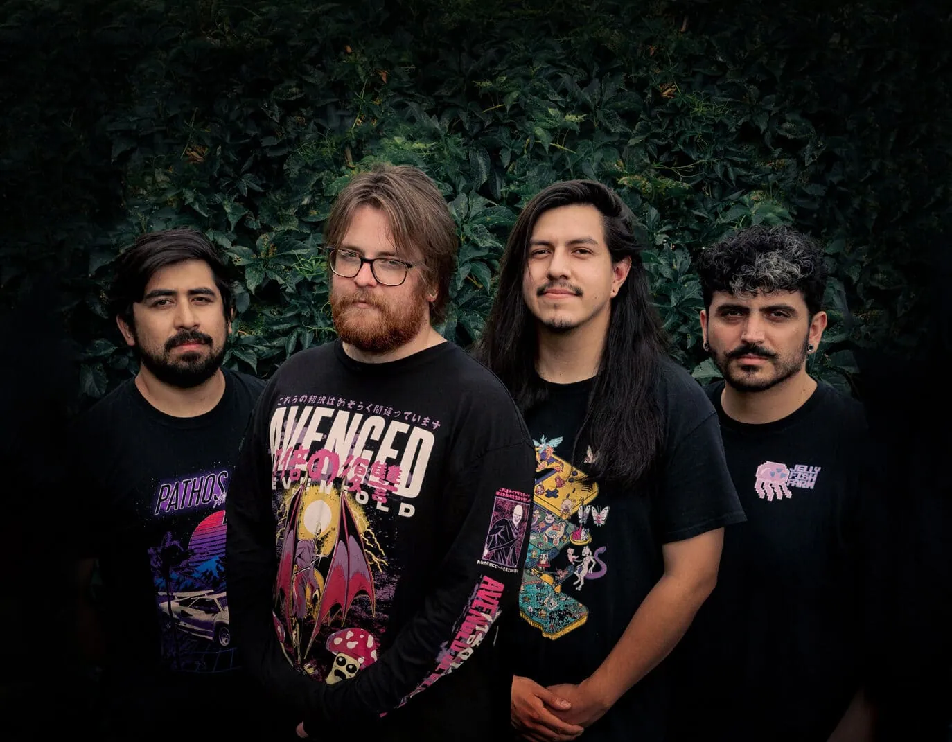 Four men in black graphic t-shirts pose in front of dense green foliage for a band portrait.