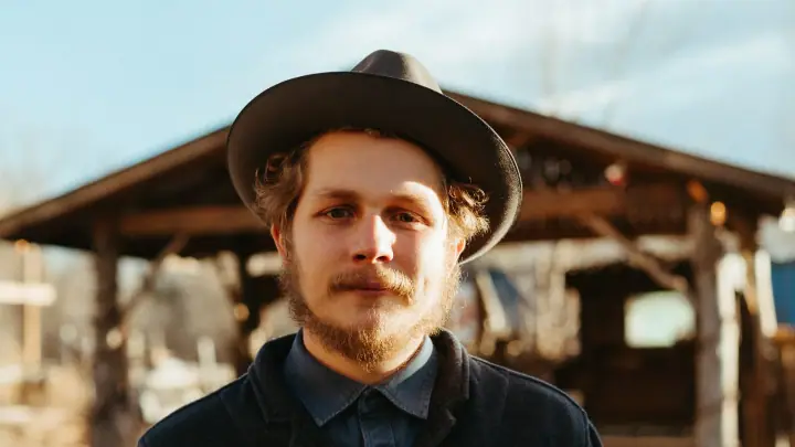 Bearded man in a dark wide-brim hat and denim-blue shirt standing before a rustic wooden pavilion on a sunny day.