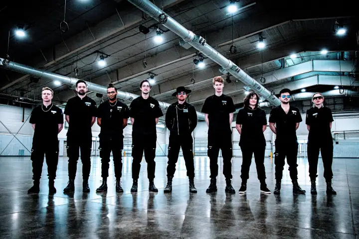 Nine people in matching black outfits stand in a row inside a large warehouse with exposed pipes and overhead lights.