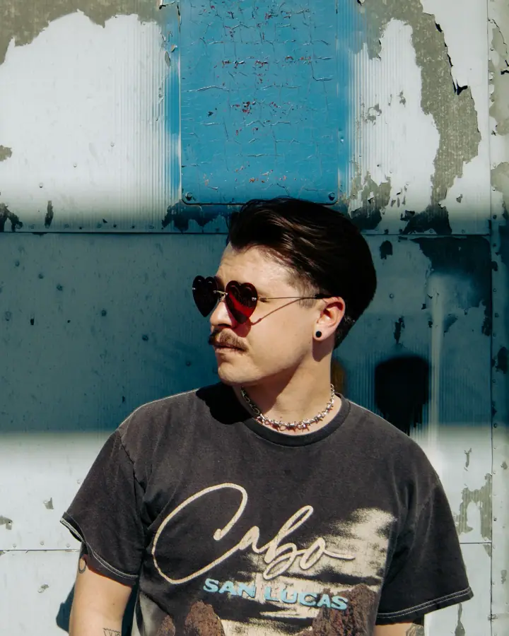 A man in heart-shaped sunglasses and a faded graphic tee stands before a peeling blue and white metal wall.