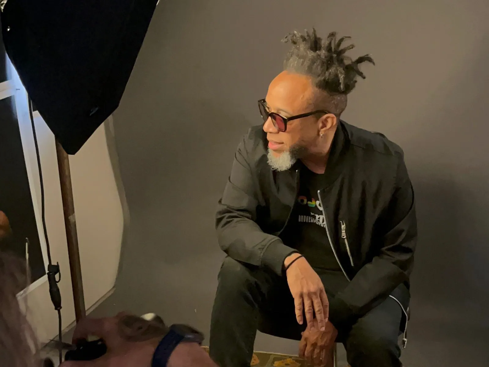 A man with gray dreadlocks and dark glasses sits in a studio beside a softbox light and gray backdrop.