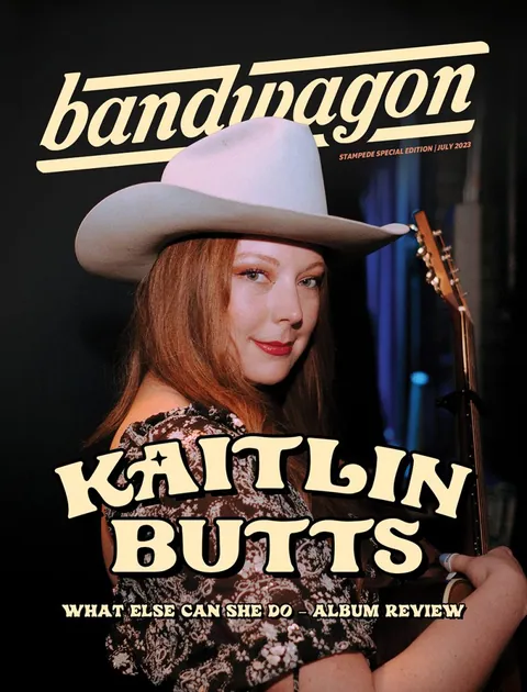 Kaitlin Butts in a cream cowboy hat and patterned blouse holds an acoustic guitar against a dark backdrop.