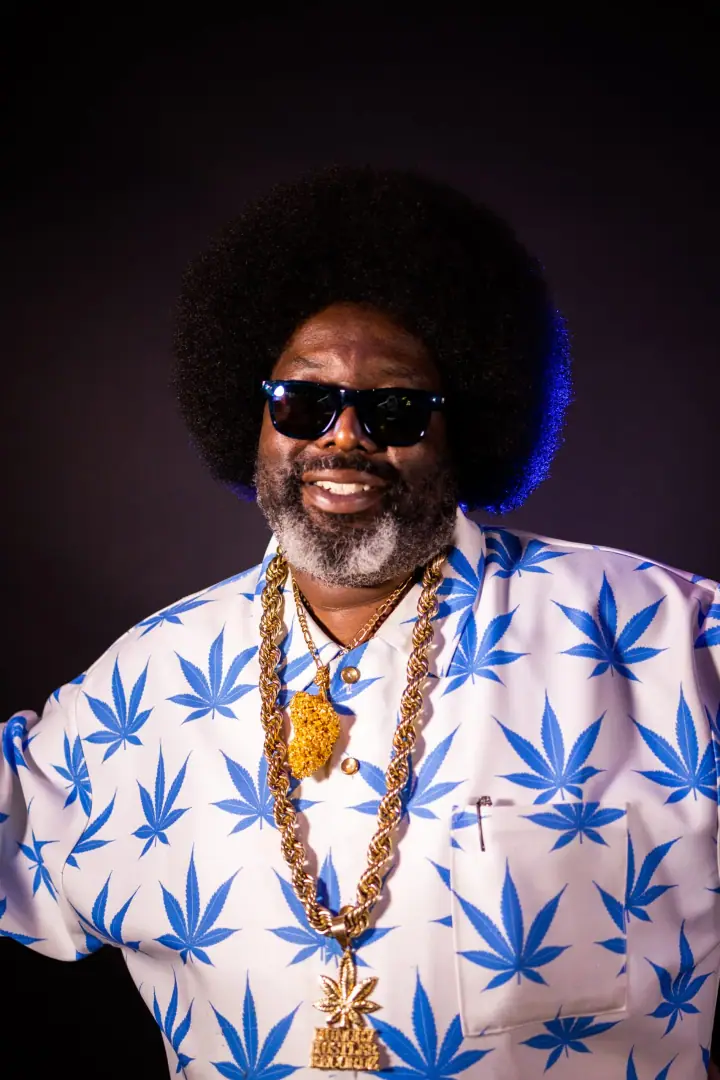 Smiling man with a big afro and sunglasses in a blue leaf-print shirt and gold chains against a dark backdrop.
