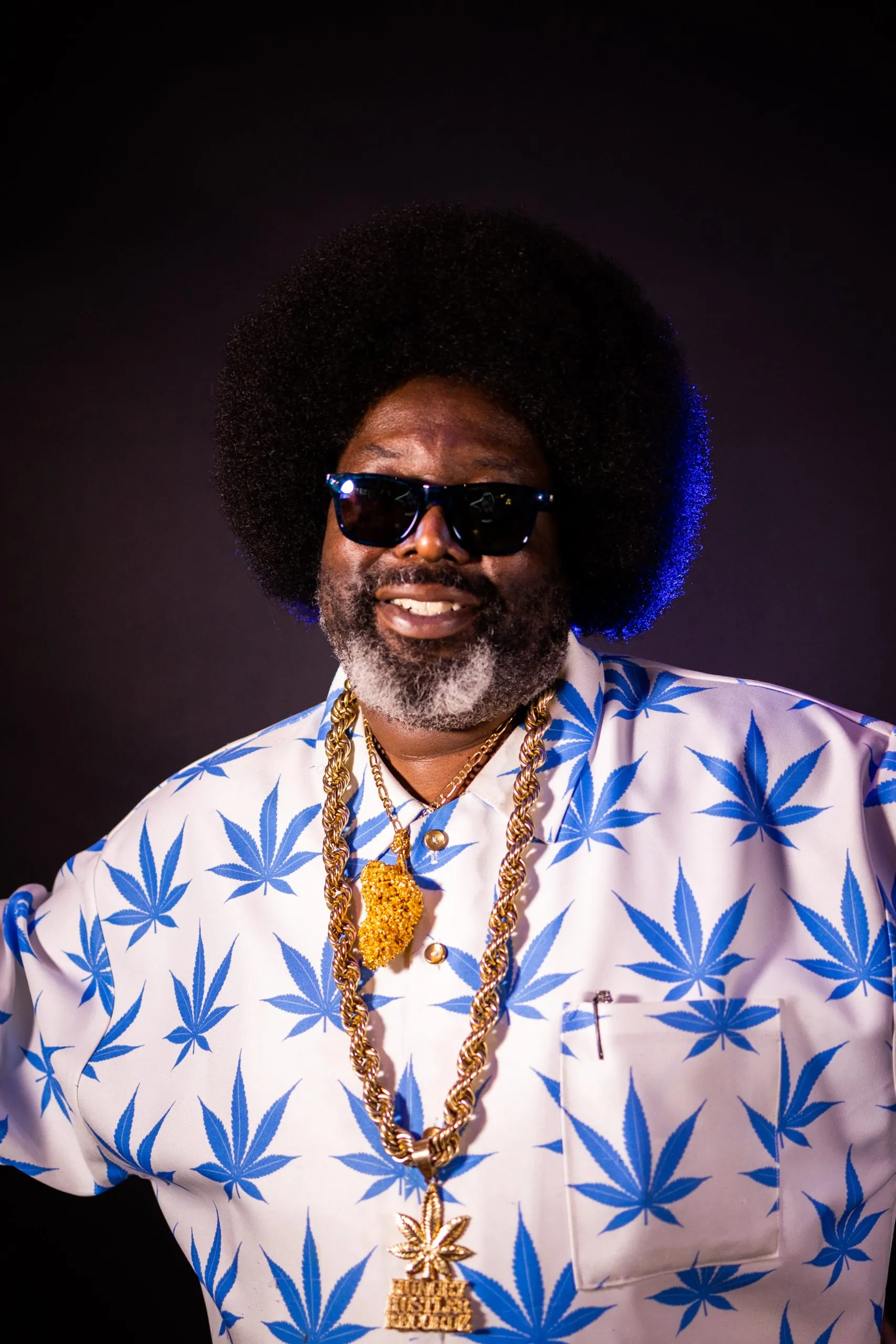 Smiling man with a big afro and sunglasses in a blue leaf-print shirt and gold chains against a dark backdrop.
