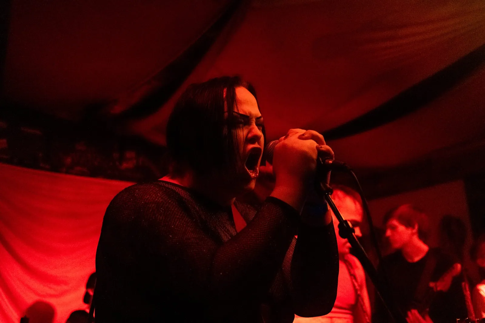 A dark-haired singer screams into a microphone under red stage lights, with musicians behind her.