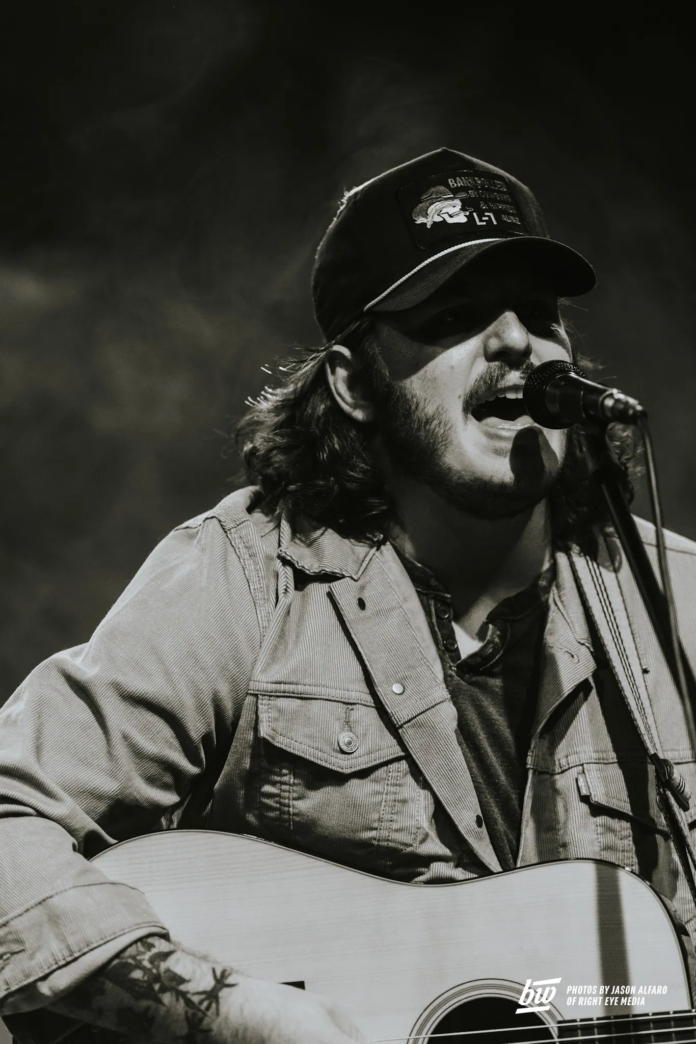 Black and white shot of a bearded man in a patched cap singing into a mic while playing acoustic guitar.