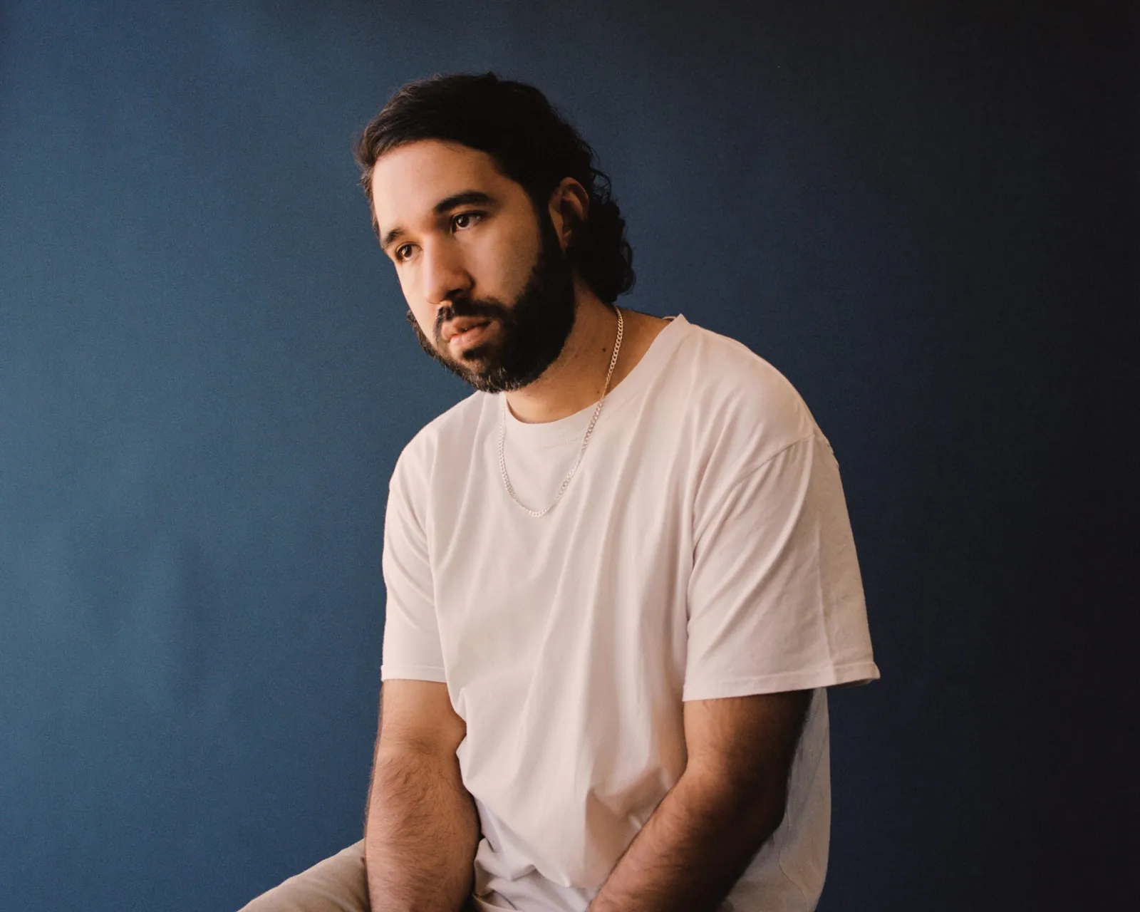 Bearded man with dark shoulder-length hair in a white t-shirt and gold chain, seated against a blue backdrop.