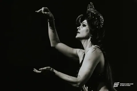 Black and white photo of a performer in a sparkly horned headdress and glitter eye makeup, arms raised on stage.
