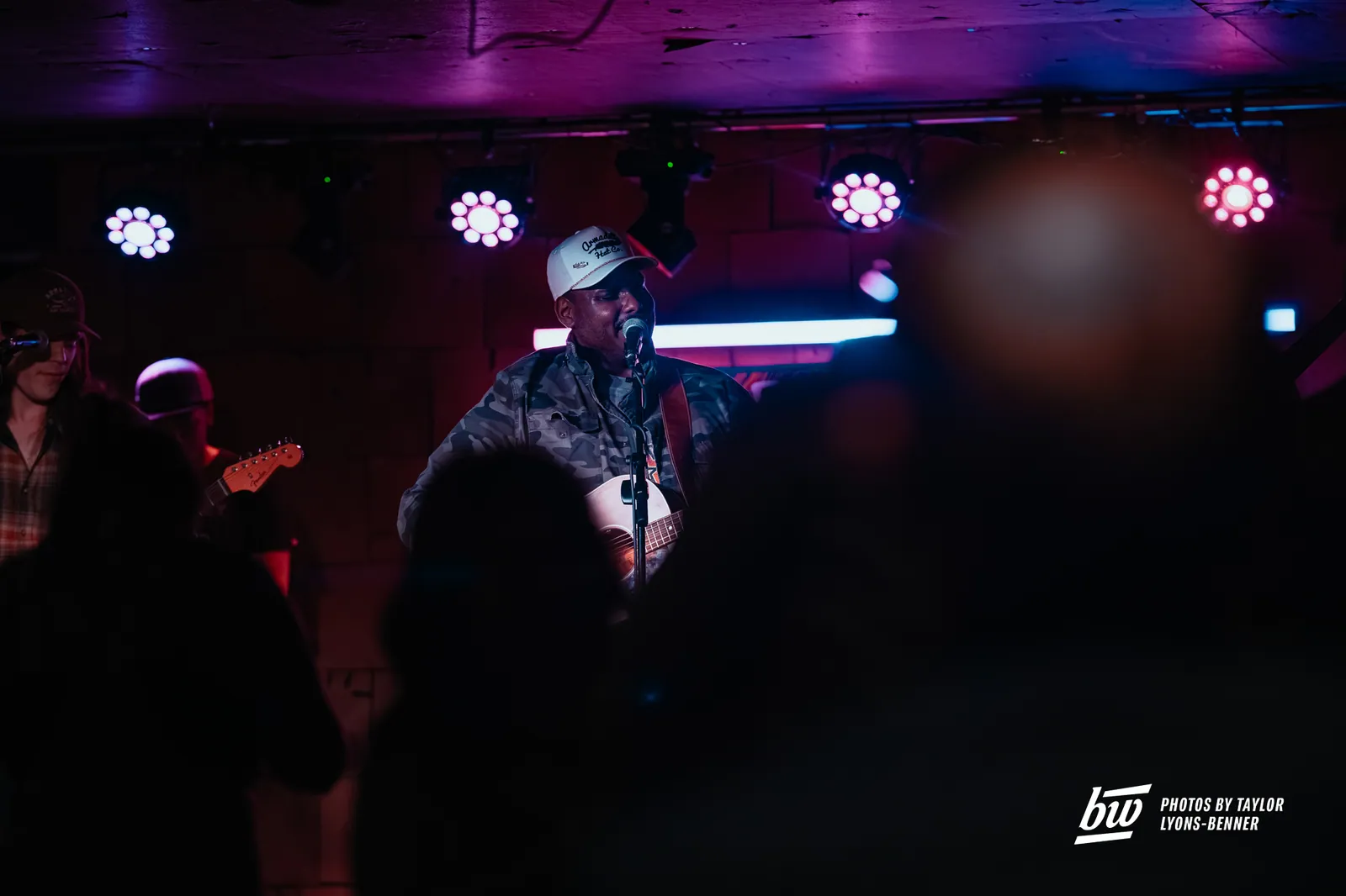 Rodell Duff performs with an acoustic guitar at a microphone under purple stage lights at The Black Buzzard.