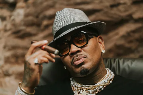 Man in a gray woven fedora and tinted sunglasses holds a cigar, wearing layered chains, before a rocky backdrop.