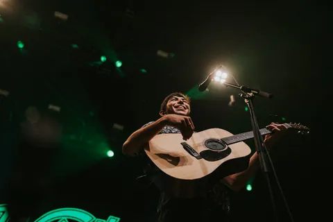 Wyatt Flores smiles while playing an acoustic guitar at a microphone under green stage lights.