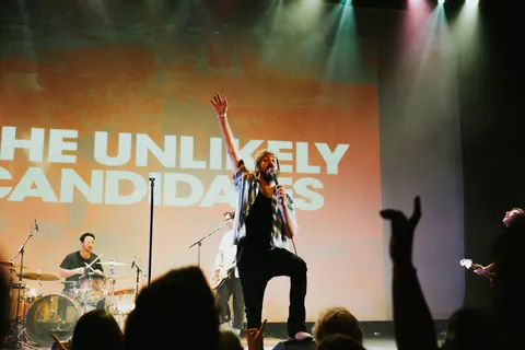 The Unlikely Candidates perform, with a singer holding a mic, a drummer, a guitarist, and raised hands in the crowd.