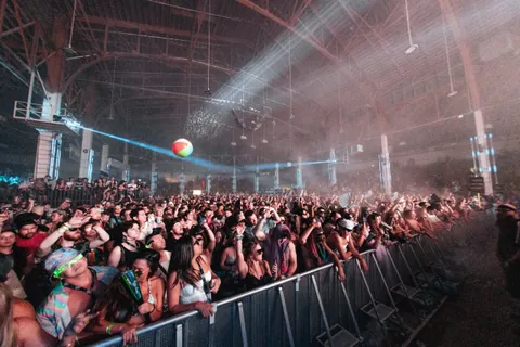 Crowd behind a metal barricade watches a beach ball float under hazy stage lights at a large indoor festival.