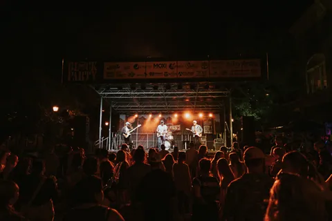 Crowd of silhouetted fans watches three musicians perform on an outdoor stage at night under warm orange lights.