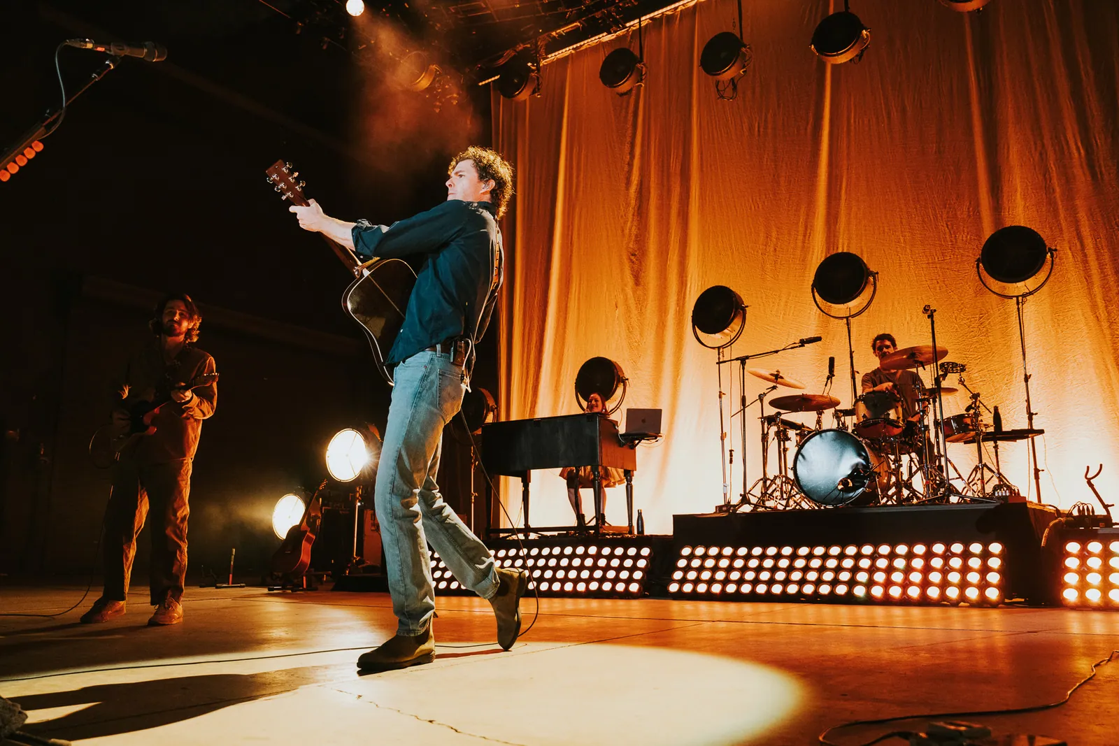 Vance Joy performs with an acoustic guitar at center stage as bandmates play under warm orange lights.