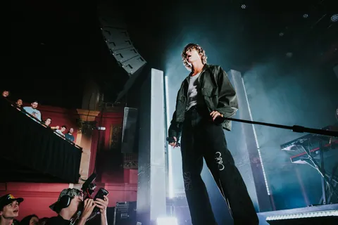 David Kushner performs on stage in a dark jacket and jeans, singing into a mic before a crowd and a photographer.