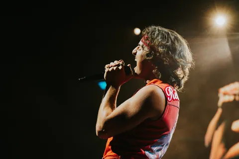 A singer in a red sleeveless top and headband grips a microphone and sings on a dark stage under a spotlight.