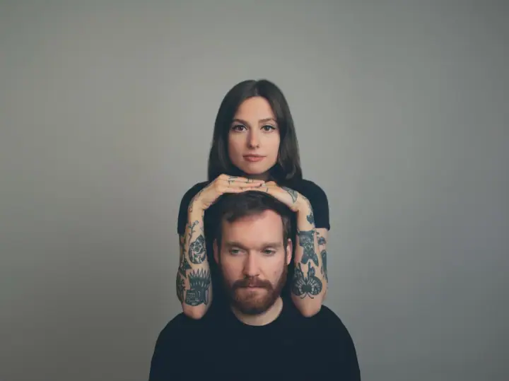 A woman with long dark hair rests her tattooed hands on the head of a bearded man in black against a gray backdrop.