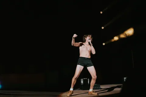 Shirtless performer in black briefs and tan shoes sings into a mic on a dark stage under spotlights.
