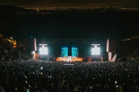 Packed crowd with raised phones faces a lit stage and large screens at a night concert in a rock amphitheater.