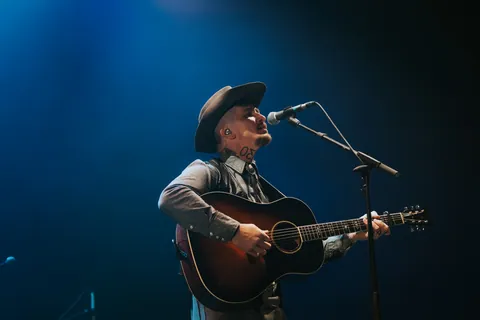 Benjamin Tod sings and plays an acoustic guitar at a microphone, wearing a brown hat under blue stage lights.