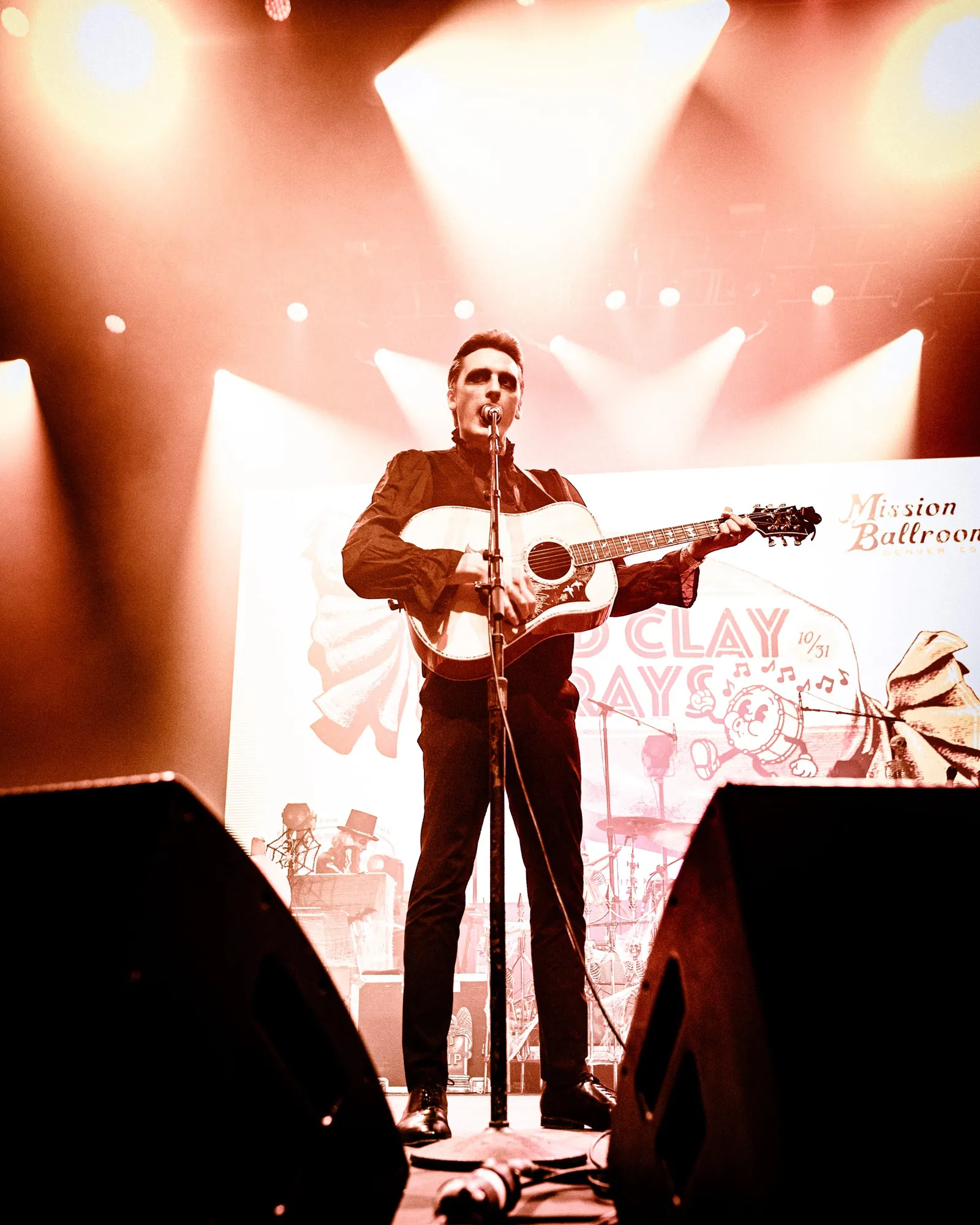 A man in a black shirt and pants sings into a microphone while playing an acoustic guitar under warm stage lights.