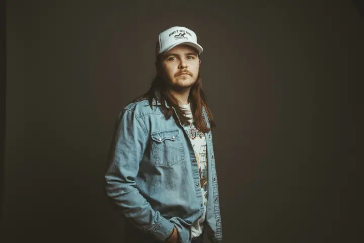A man with long brown hair and a mustache wears a white cap and denim shirt against a dark backdrop.