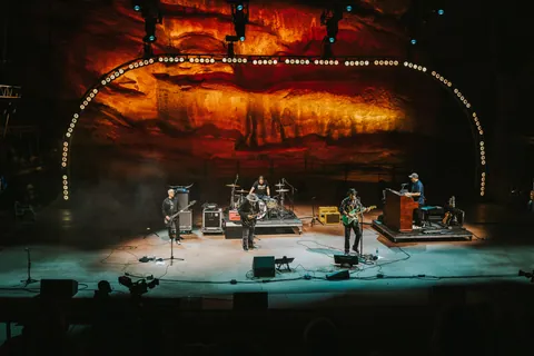 Big Head Todd and the Monsters perform with five musicians on stage before a glowing red rock backdrop at Red Rocks.