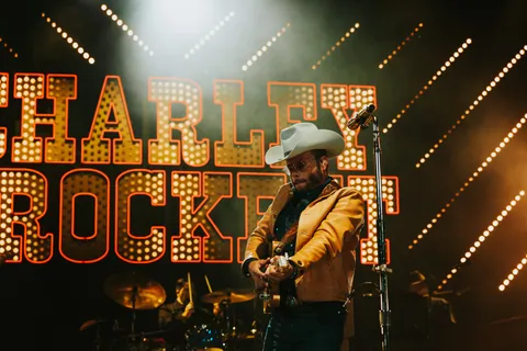 Charley Crockett performs on stage in a white cowboy hat and gold jacket beneath a marquee sign.