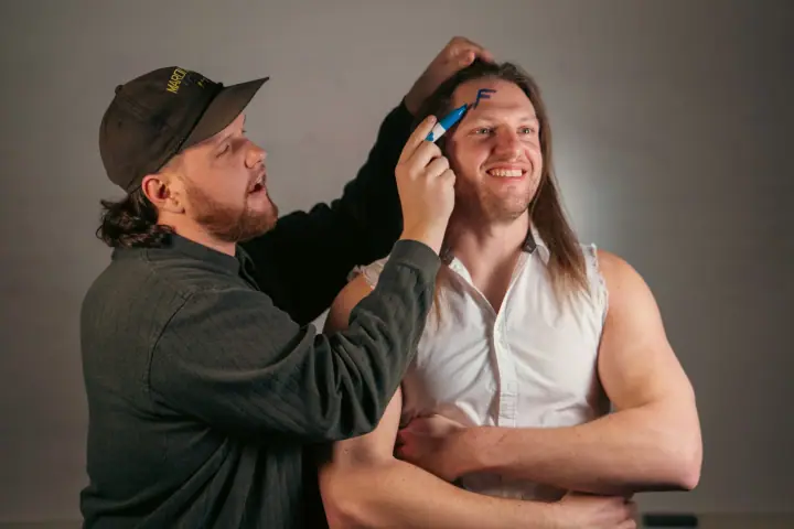 A bearded man in a cap draws on the forehead of a smiling long-haired man in a sleeveless shirt with a blue marker.