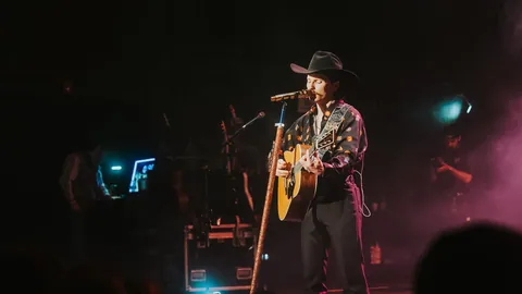 Cowboy-hatted performer in a patterned shirt sings and strums an acoustic guitar under purple stage lights.