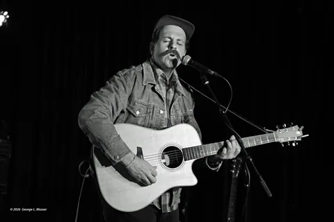 Black-and-white photo of a mustached singer in a cap and denim jacket playing an acoustic guitar at a microphone.
