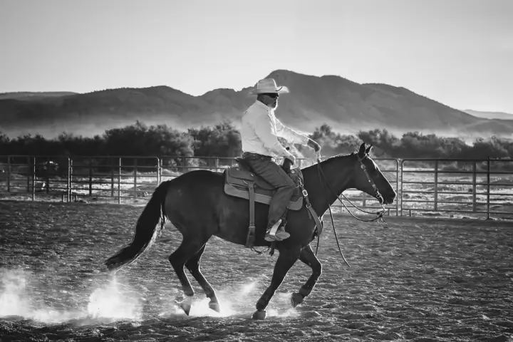 Black and white photo of a cowboy in a hat and white shirt riding a dark horse in a dusty arena.