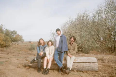 Prairie's four members pose on a weathered log in a dry field, with trees and an overcast sky behind them.
