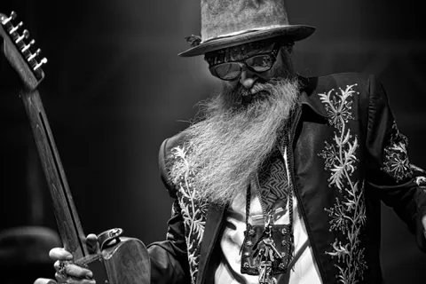 Black-and-white photo of a bearded musician in a wide-brimmed hat playing guitar on a dark stage.