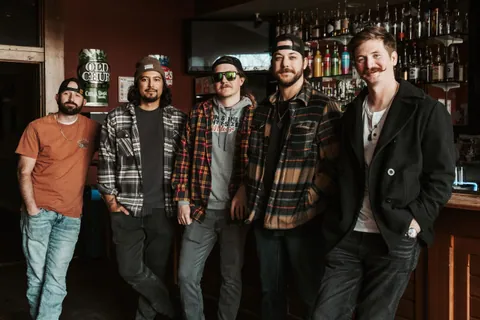 Five men in flannel shirts, caps, and a peacoat pose together in a bar in front of shelves of liquor bottles.