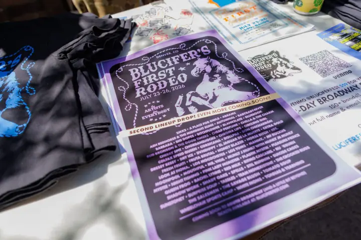 A table shows a Blucifer's First Rodeo lineup poster, black horse-print t-shirts, and promotional stickers.