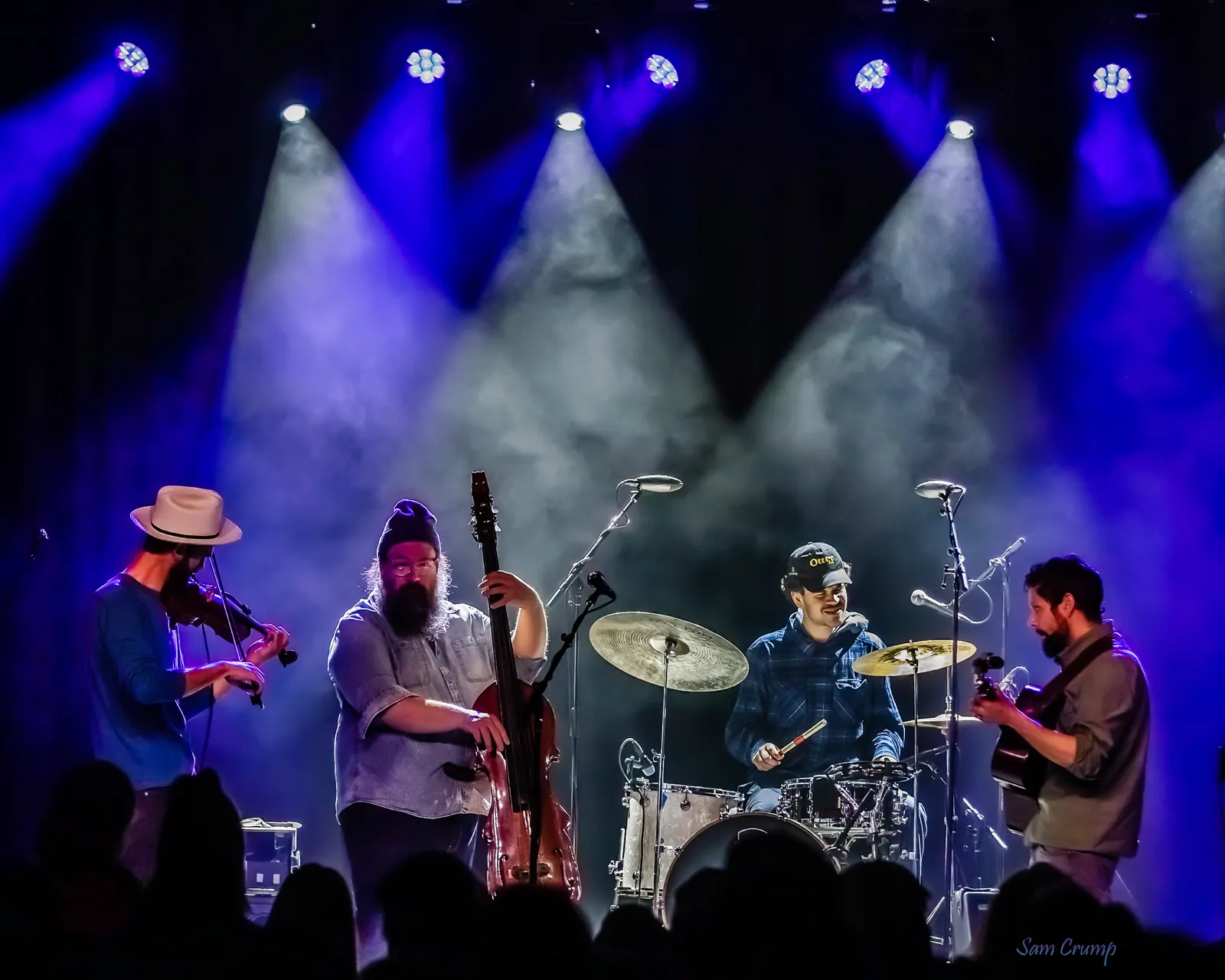 Four musicians play fiddle, upright bass, drums, and guitar on a smoky, blue-lit stage.