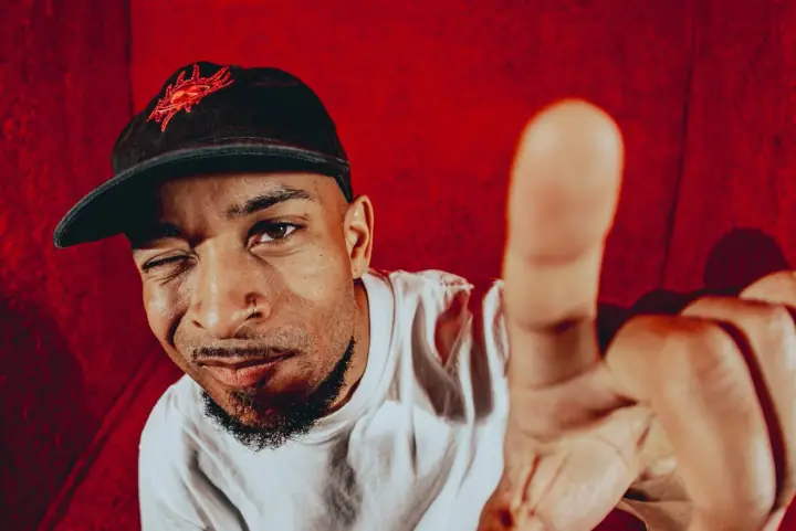 A man in a black cap with a red embroidered logo winks and points toward the camera against a red textured backdrop.