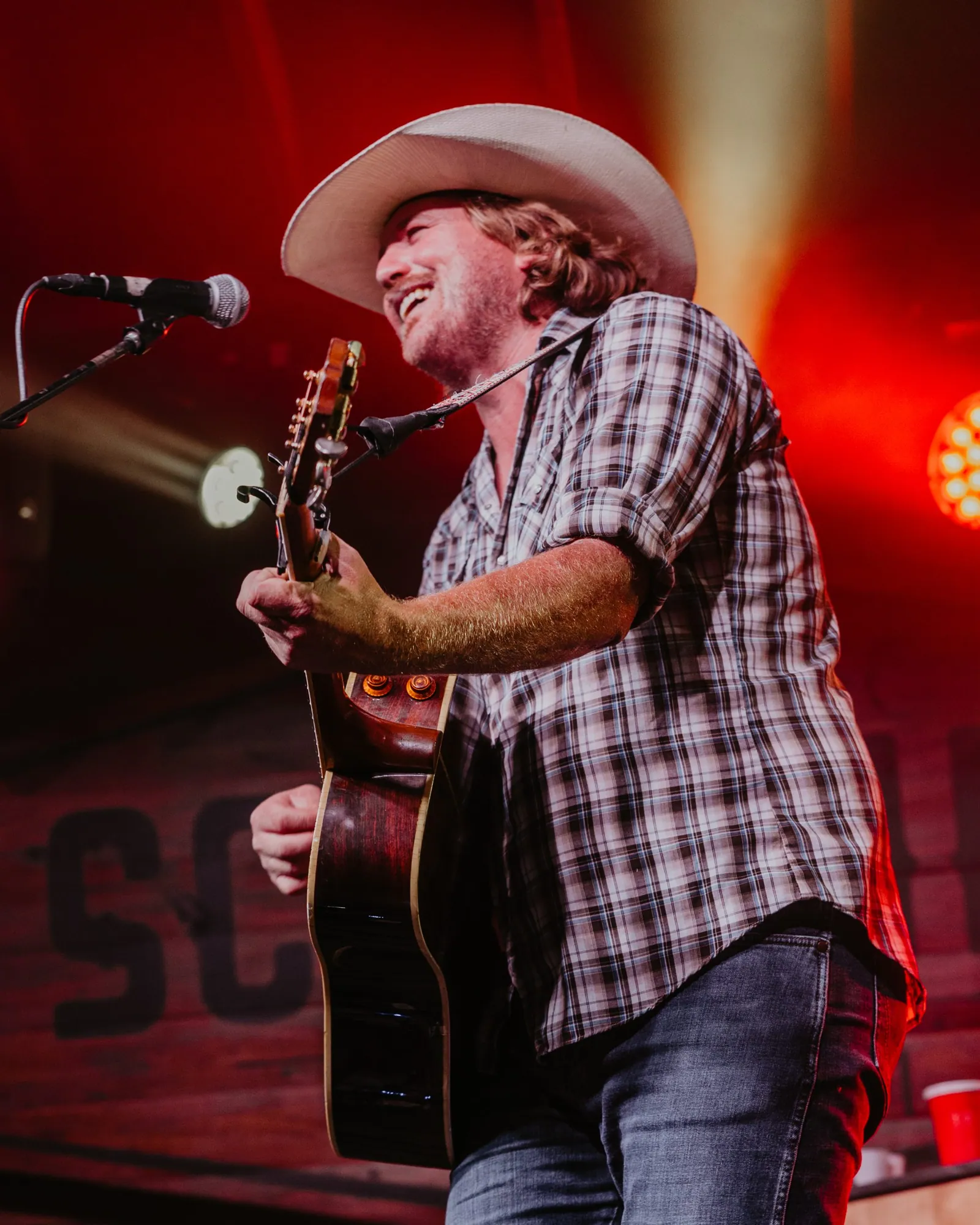 A smiling man in a cream cowboy hat and plaid shirt plays acoustic guitar at a mic under red stage lights.