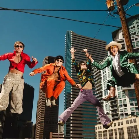 Four people in bright retro outfits leap midair on a rooftop with skyscrapers and power lines behind them.