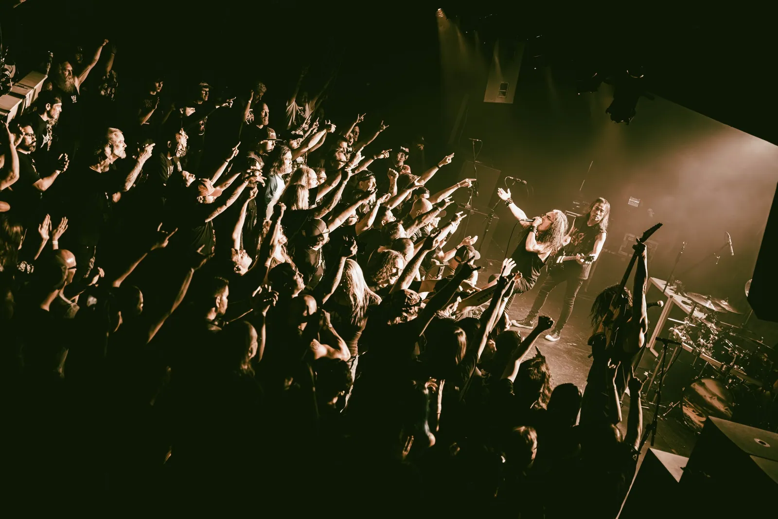 Death Angel performs at the Moxi Theater as fans with raised arms crowd the stage under amber lights.