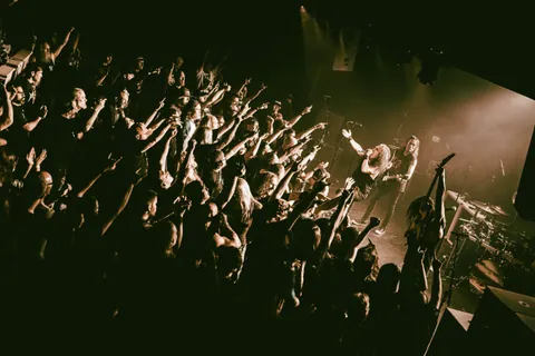 Death Angel performs at the Moxi Theater as fans with raised arms crowd the stage under amber lights.