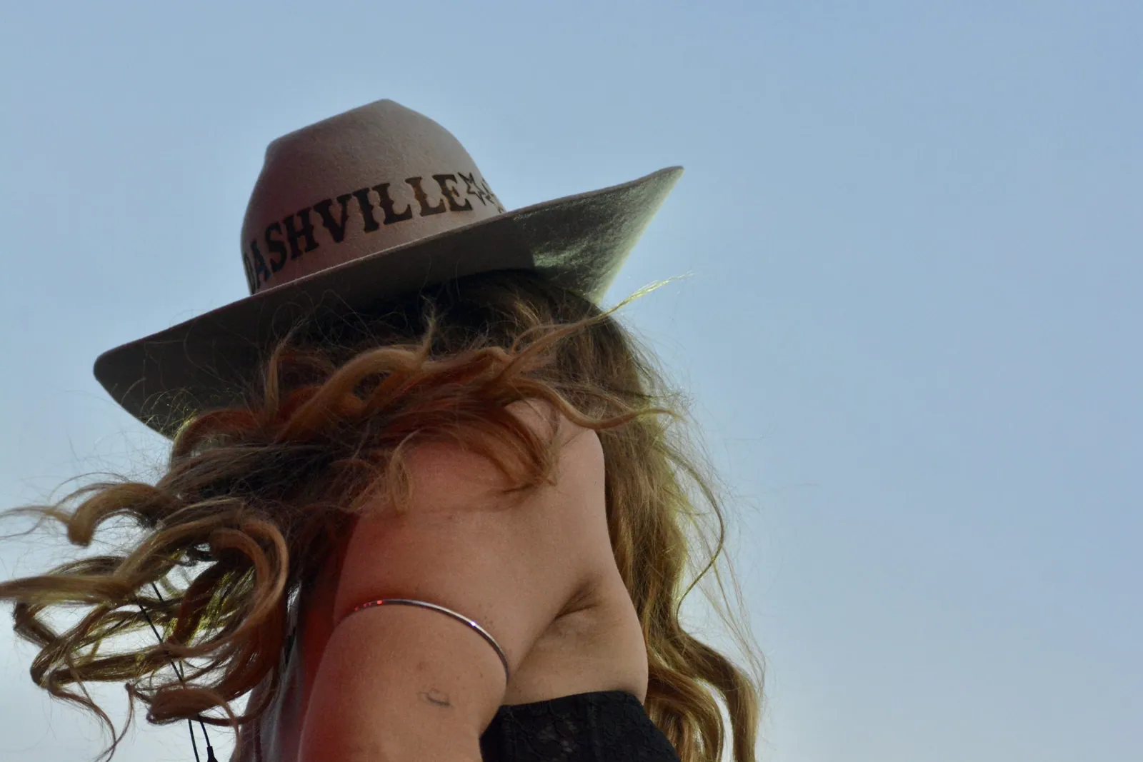 A woman with wind-blown auburn curls wears a gray felt hat with "Nashville" on the band against a pale blue sky.