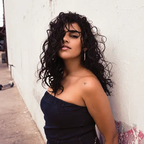 A woman with long dark curly hair leans against a white wall in a strapless denim top and a drop earring.