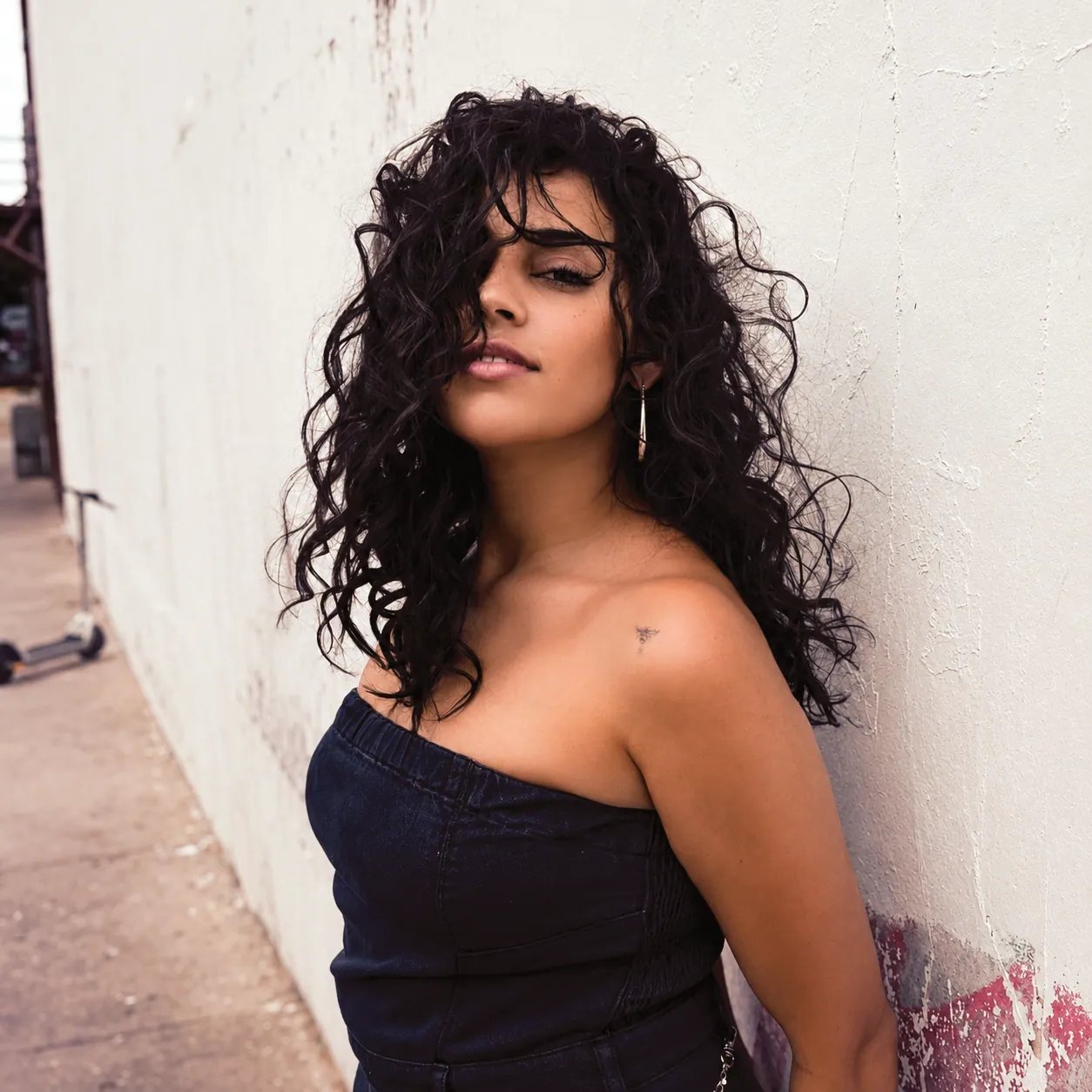 A woman with long dark curly hair leans against a white wall in a strapless denim top and a drop earring.