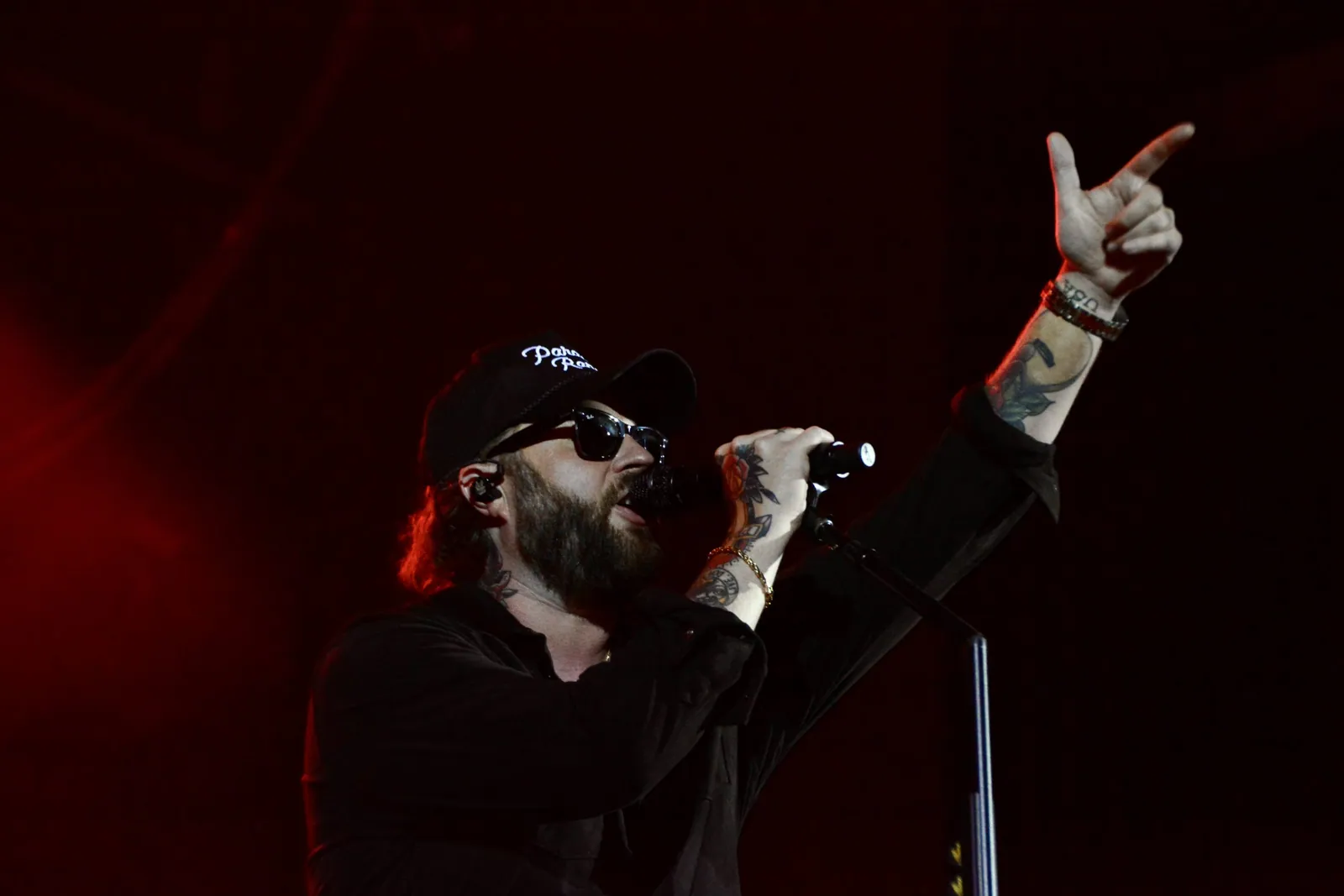 A bearded performer in a black cap and sunglasses sings into a mic while pointing up under red stage lights.